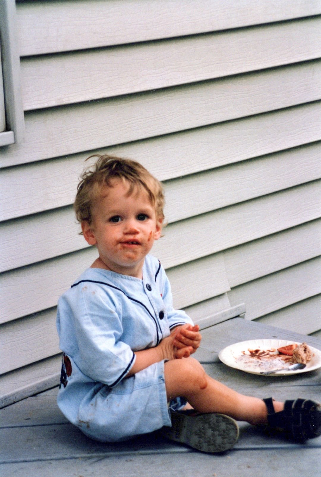 A toddler in a light blue outfit sitting on the back porch, chocolate on his face, an almost-finished treat on a paper plate beside him