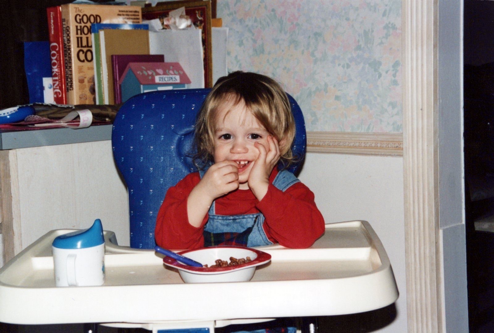 A toddler in denim overalls grinning in a high chair over a bowl of cereal, cookbooks on the shelf behind